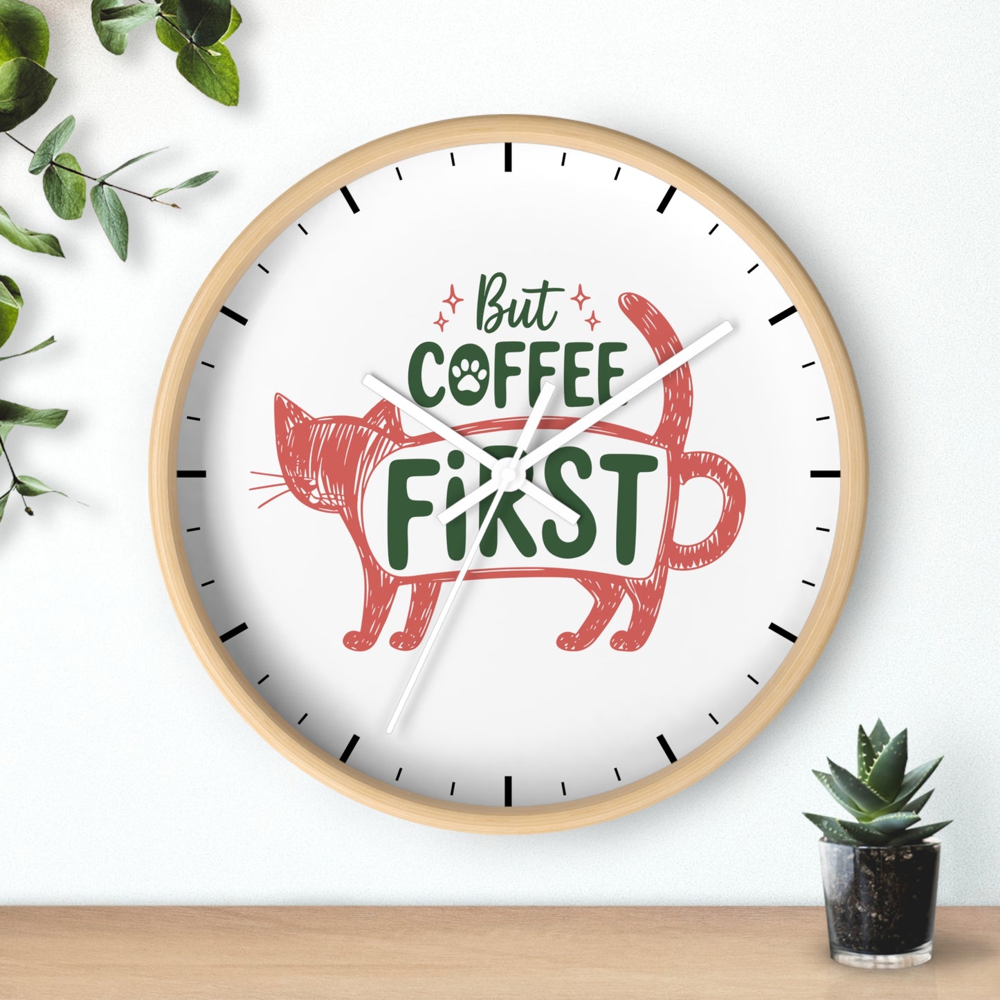 A round wall clock with a light wood frame and a white face, featuring the phrase 'But COFFEE FIRST' in green and red lettering with a red cat illustration. The clock is mounted on a white wall above a wooden surface, with decorative green leaves and a small potted succulent nearby.