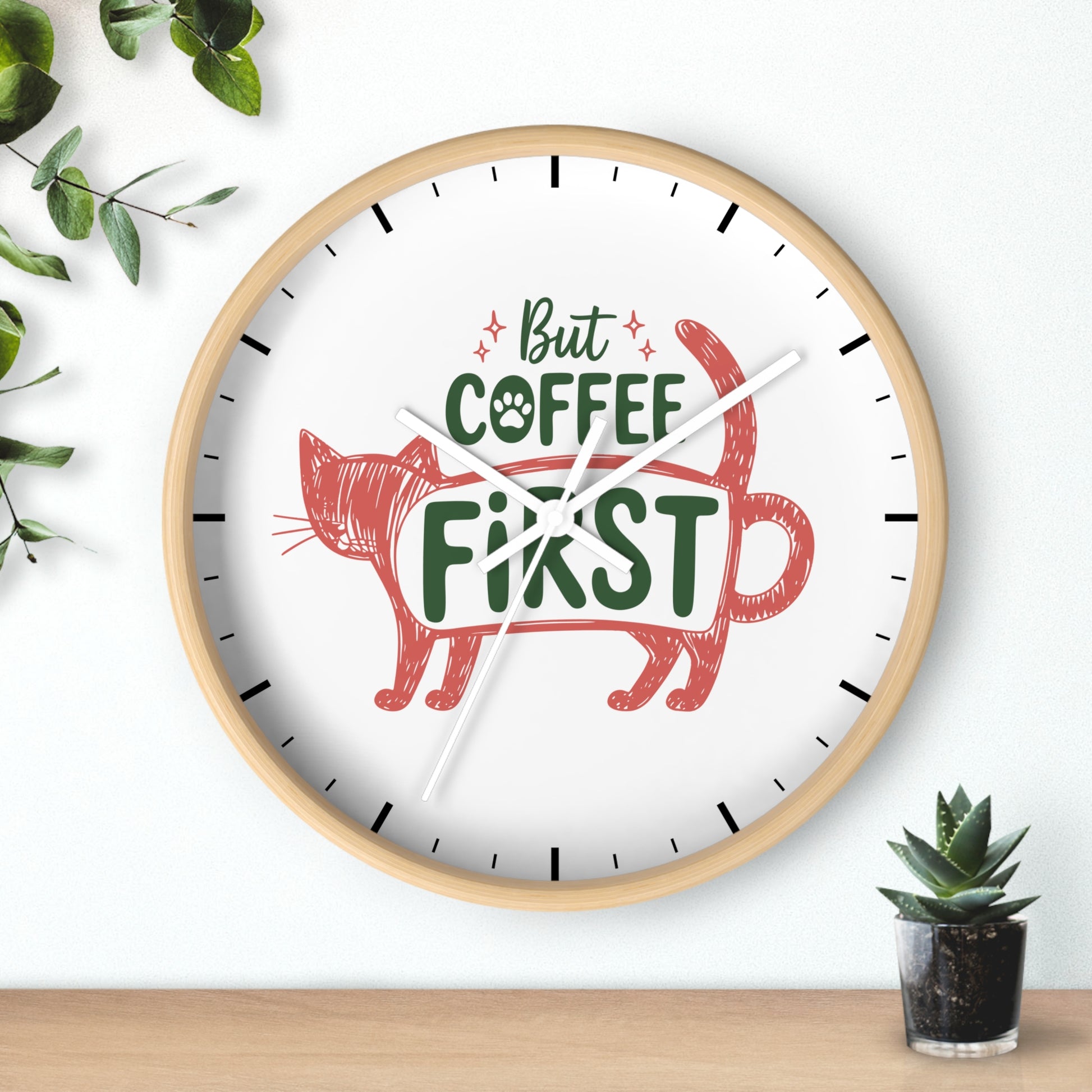 A round wall clock with a light wood frame and a white face, featuring the phrase 'But COFFEE FIRST' in green and red lettering with a red cat illustration. The clock is mounted on a white wall above a wooden surface, with decorative green leaves and a small potted succulent nearby.