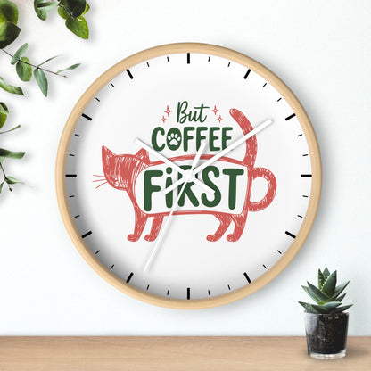 A round wall clock with a light wood frame and a white face, featuring the phrase 'But COFFEE FIRST' in green and red lettering with a red cat illustration. The clock is mounted on a white wall above a wooden surface, with decorative green leaves and a small potted succulent nearby.