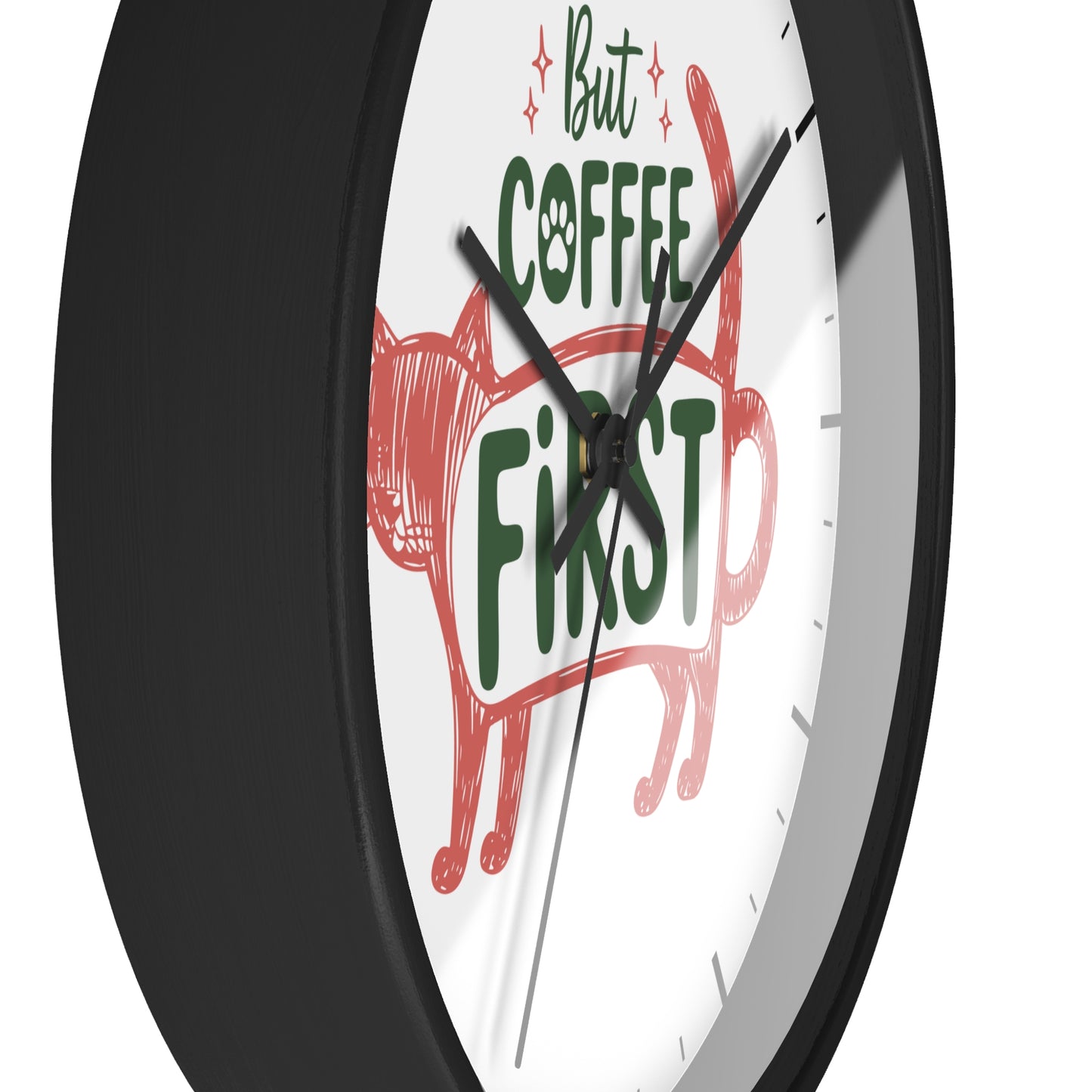 A close-up of a round wall clock with a black frame and white face, featuring the phrase 'But COFFEE FIRST' in green and red lettering with a red cat illustration.