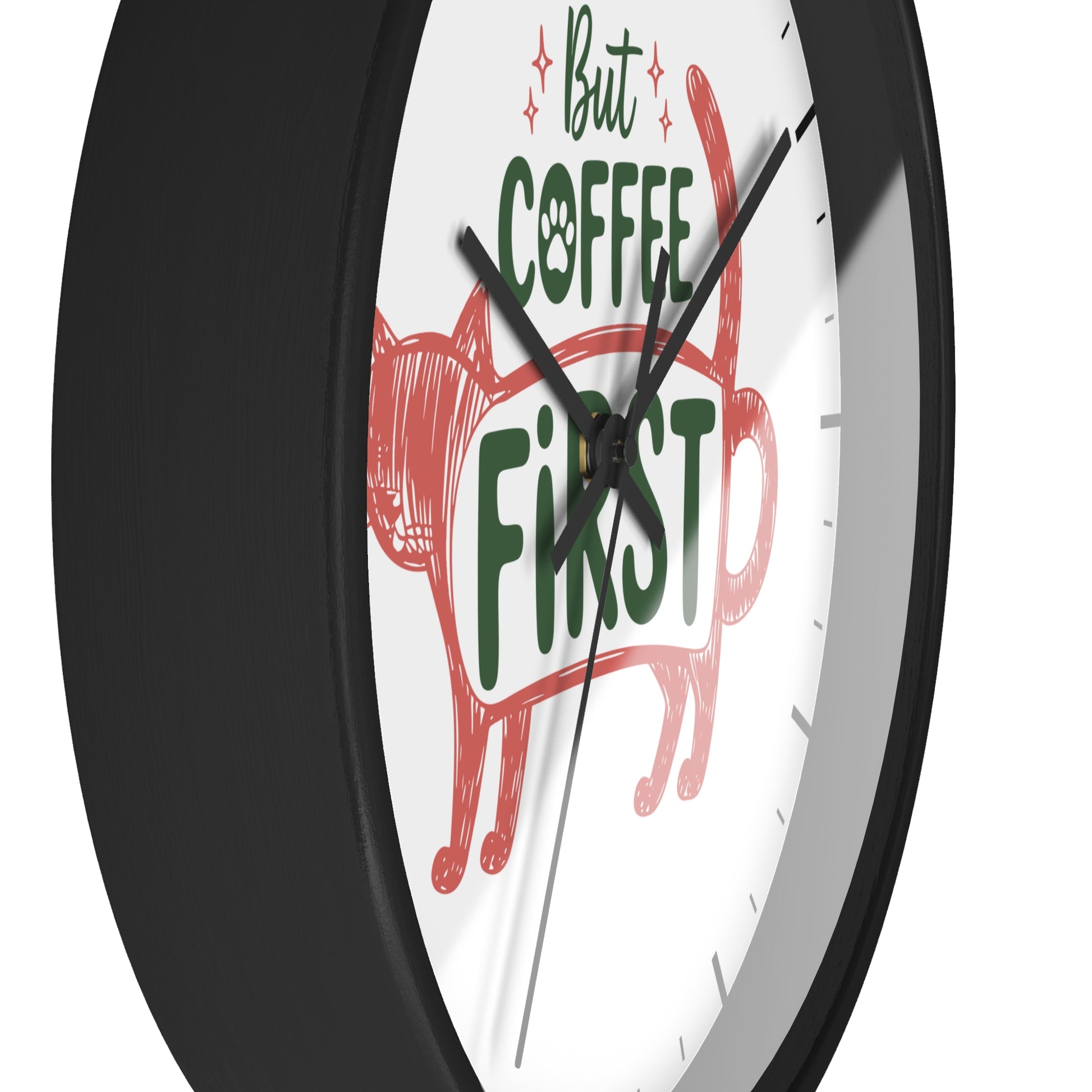 A close-up of a round wall clock with a black frame and white face, featuring the phrase 'But COFFEE FIRST' in green and red lettering with a red cat illustration.