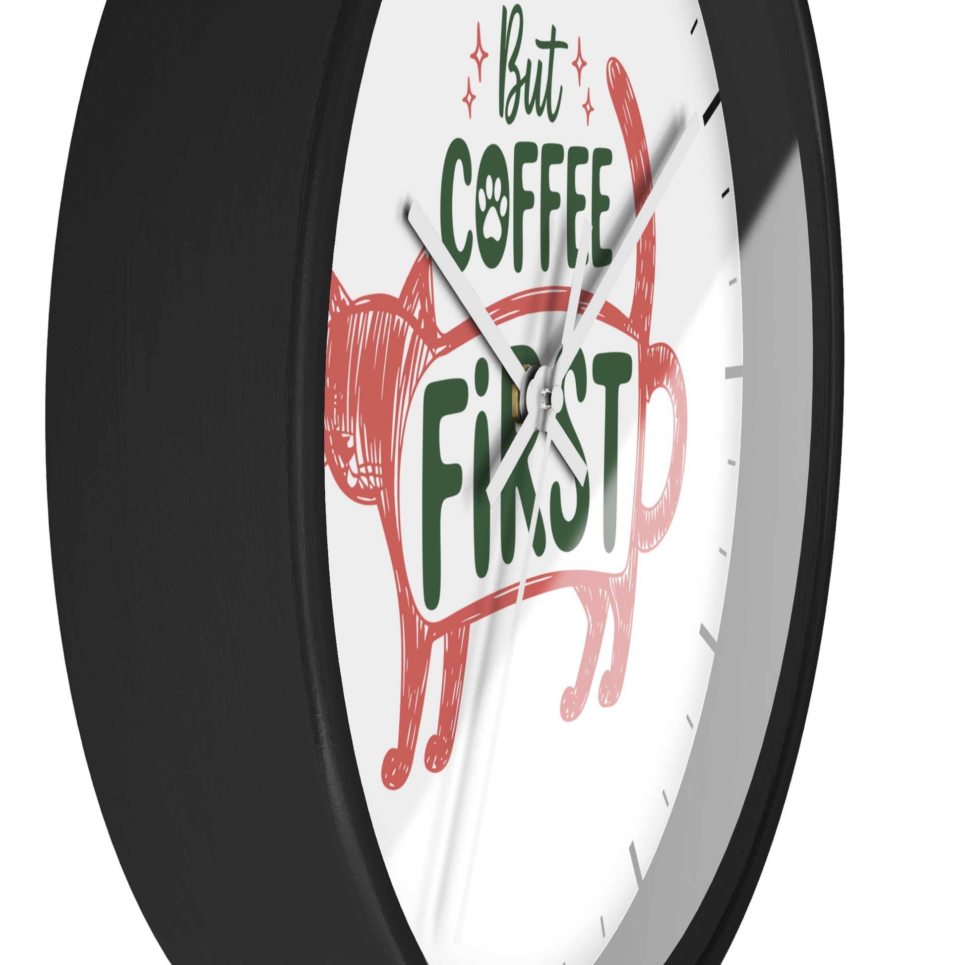 A close-up of a round wall clock with a black frame and white face. The clock face features a red cat illustration and the phrase 'But COFFEE FIRST' in green and red lettering.