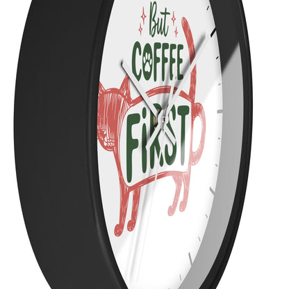 A close-up of a round wall clock with a black frame and white face. The clock face features a red cat illustration and the phrase 'But COFFEE FIRST' in green and red lettering.