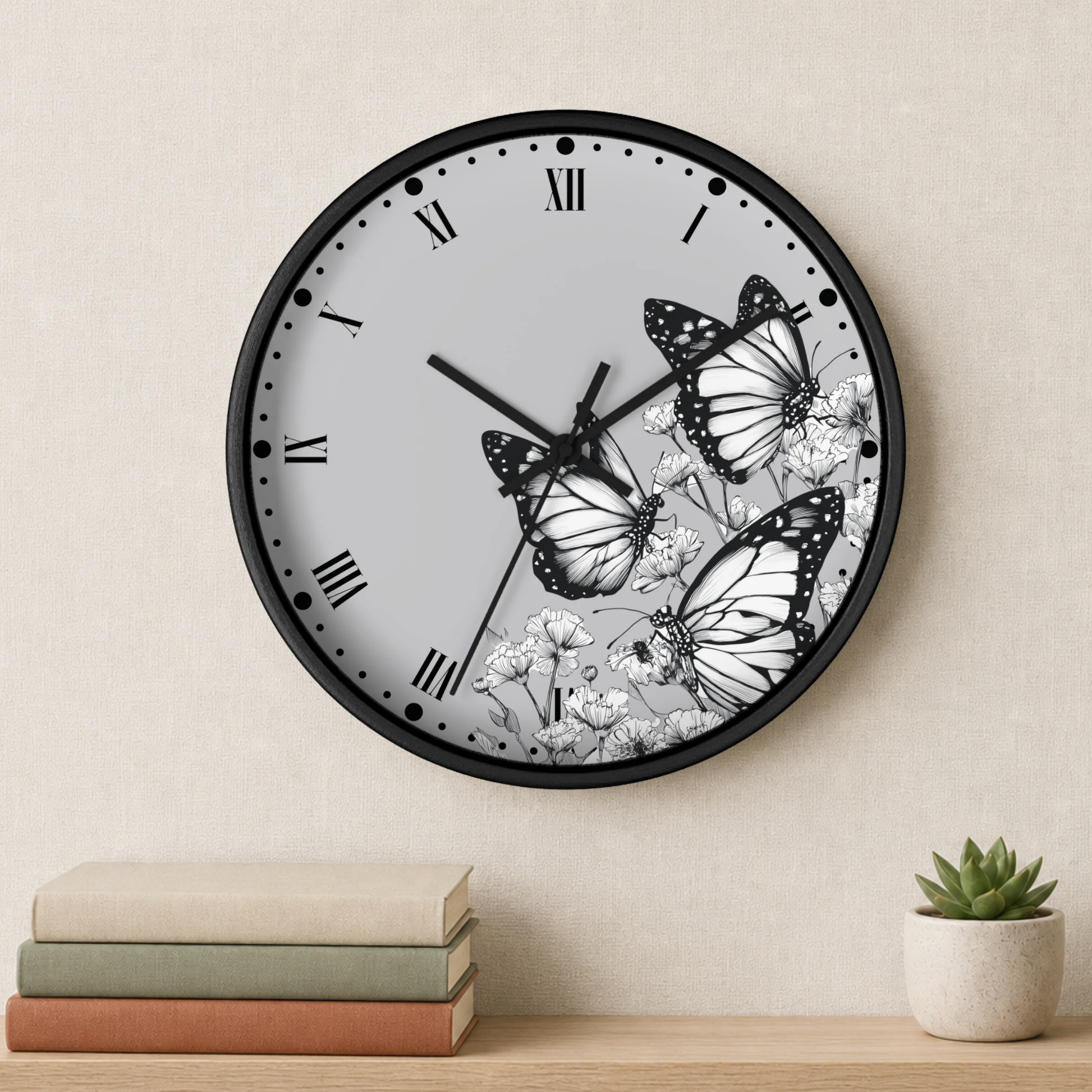 A round wall clock with a black frame and a white face featuring black-and-white butterfly and floral illustrations, mounted on a beige wall above a shelf with books and a small succulent.