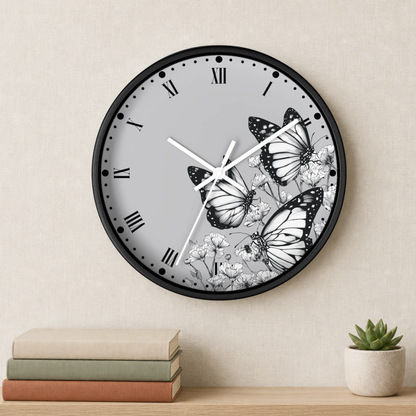 A round wall clock with a black frame and a light gray face featuring three black-and-white butterflies and small white flowers. The clock is mounted on a beige wall above a wooden shelf with stacked books and a small potted succulent.