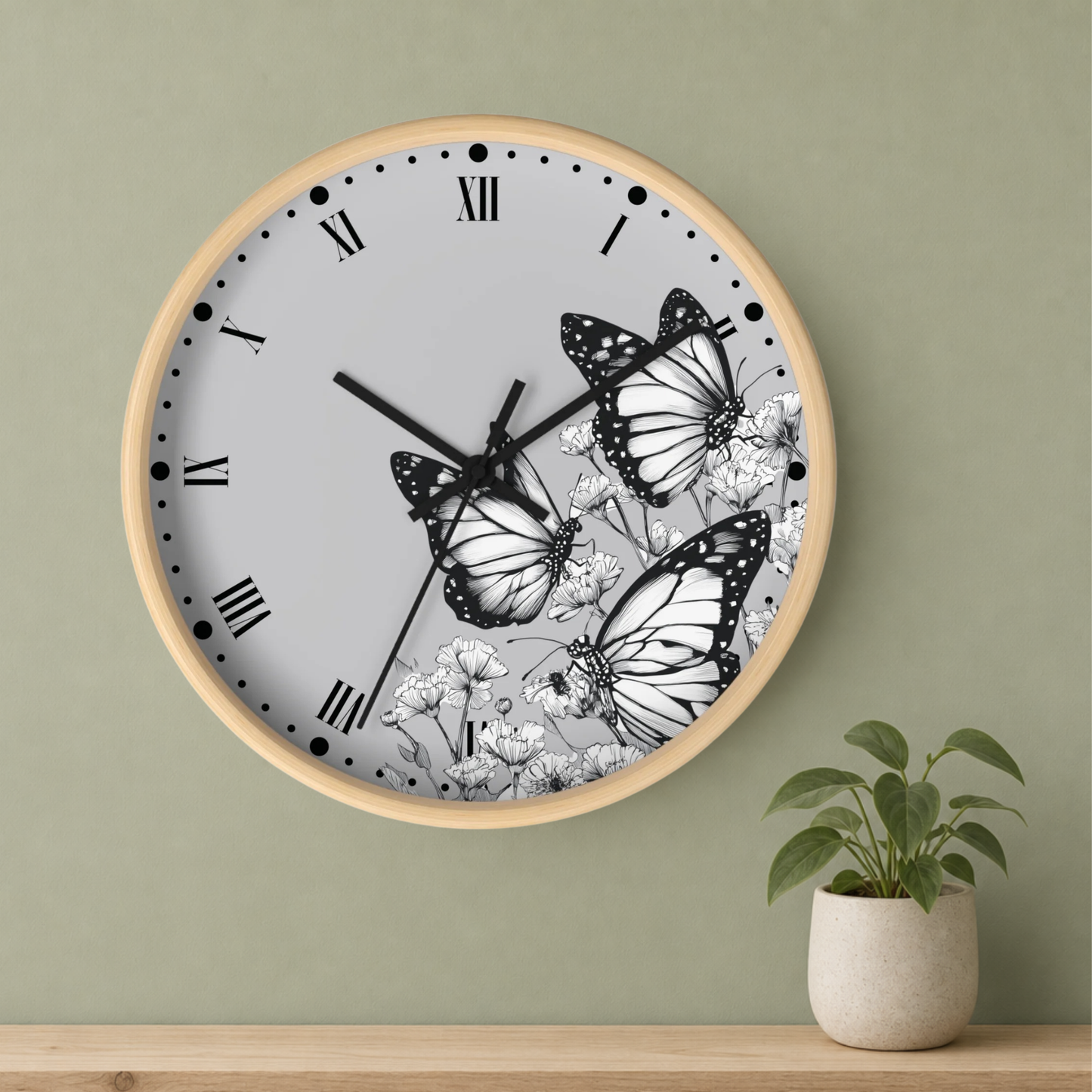 A round wall clock with a light wood frame, black Roman numerals, and a white face decorated with black-and-white butterfly and floral illustrations, mounted on a sage green wall above a wooden shelf with a small potted plant.