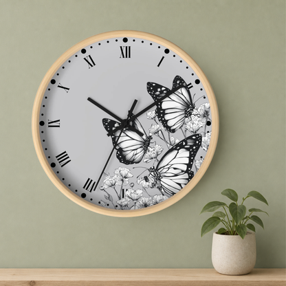 A round wall clock with a light wood frame, black Roman numerals, and a white face decorated with black-and-white butterfly and floral illustrations, mounted on a sage green wall above a wooden shelf with a small potted plant.