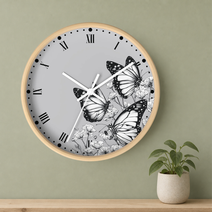 A round wall clock with a light wood frame, a gray face, black Roman numerals, and black-and-white butterfly and floral artwork, mounted on a sage green wall above a wooden shelf with a small potted plant.