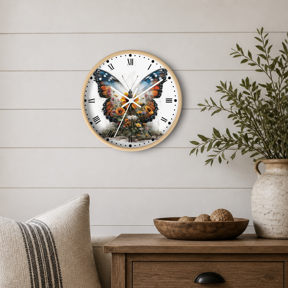 A round wall clock with a light wood frame and a detailed butterfly illustration on the face, mounted on a white shiplap wall above a wooden side table with a vase and bowl.