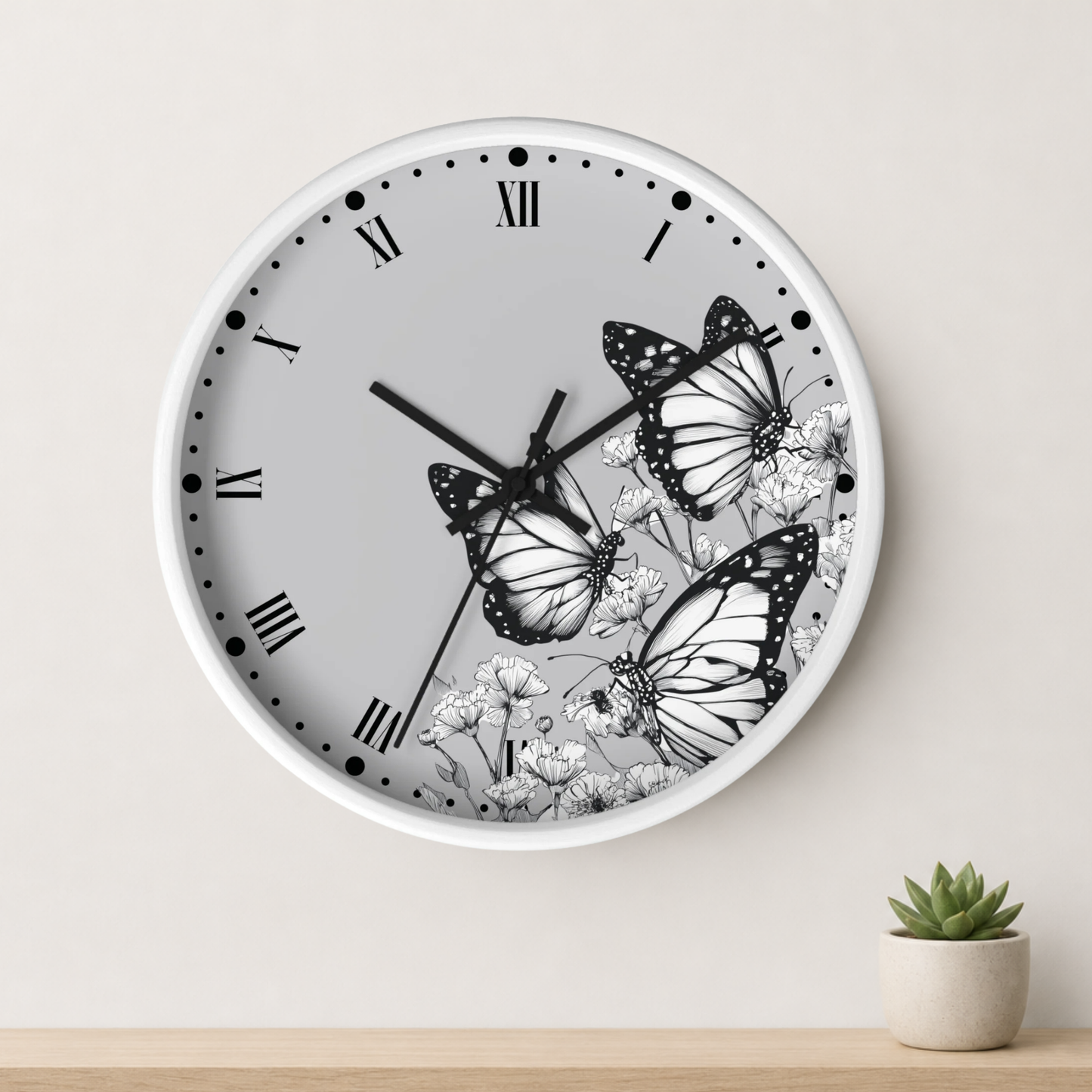 A round wall clock with a white frame and a light gray face featuring black Roman numerals and three black-and-white butterfly illustrations. The clock is mounted on a light beige wall above a wooden surface with a small potted succulent.