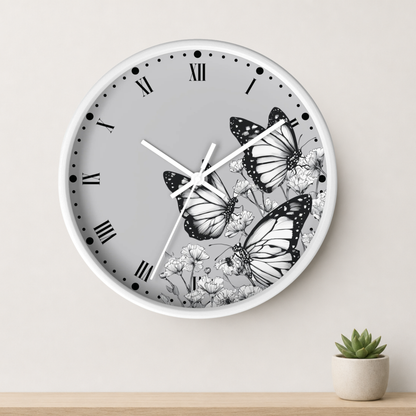 A round wall clock with a white frame and a gray face featuring black Roman numerals and butterfly illustrations, shown hanging on a light wall above a small potted succulent.