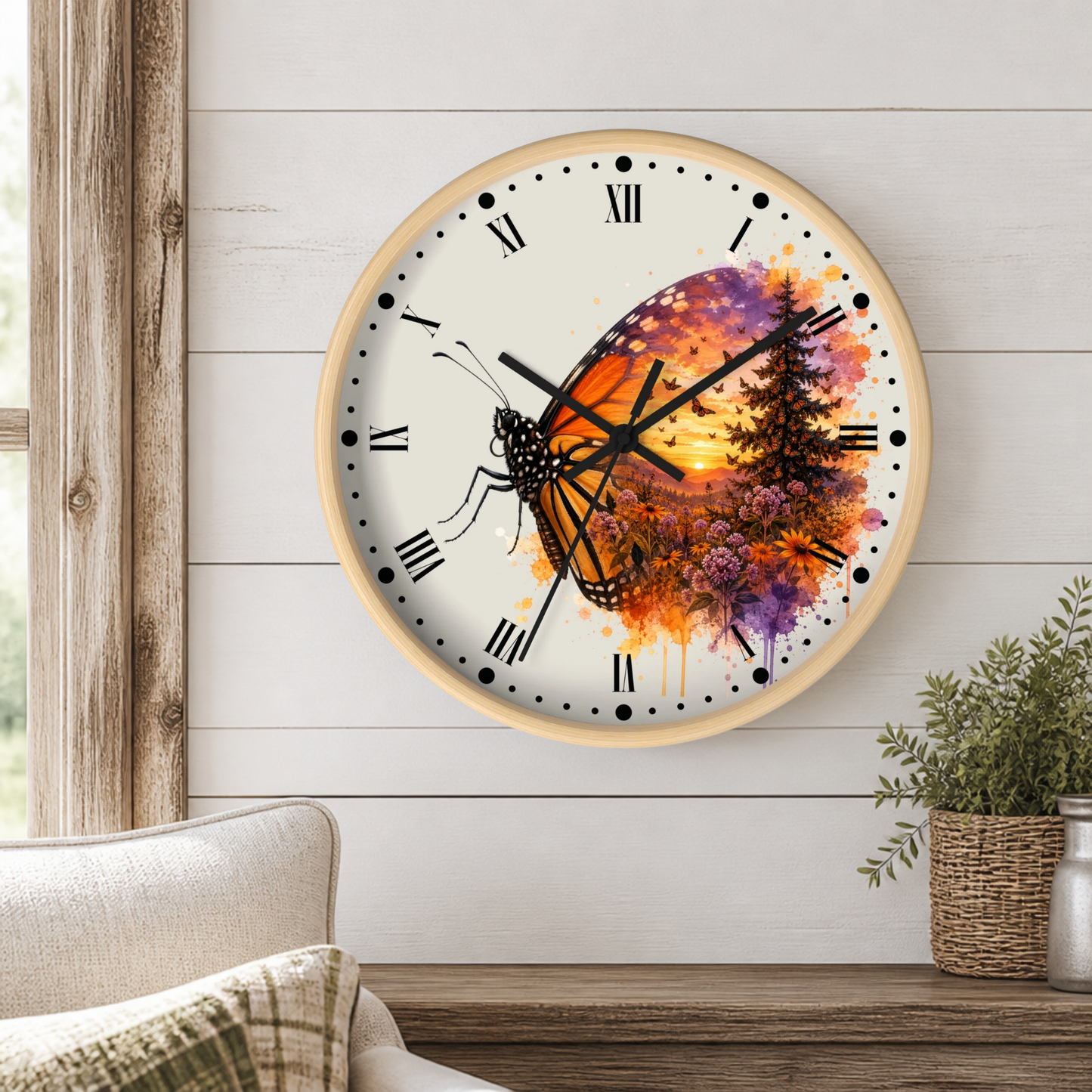A round wall clock with a light wood frame hangs on a white shiplap wall above a wooden table. The clock face features a colorful butterfly and tree design with a sunset gradient in orange, purple, and yellow on a white background with black Roman numerals.