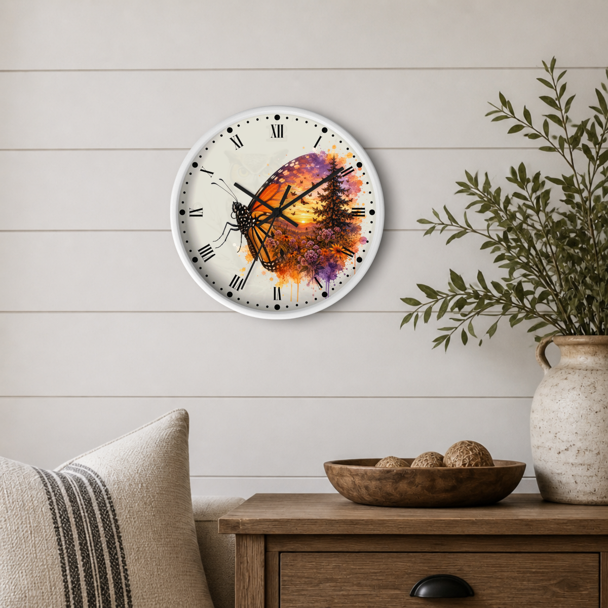 A round wall clock with a white frame and a colorful butterfly design on the face, mounted on a light beige shiplap wall above a wooden side table with a plant and decorative bowl.