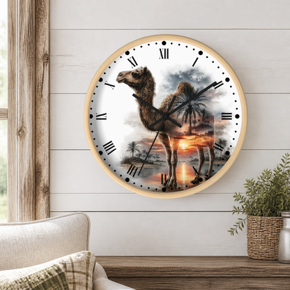 A round wall clock with a light wood frame hangs on a white shiplap wall above a wooden table. The clock face features a scenic desert landscape with a camel walking near palm trees at sunset, with black Roman numerals and hands.