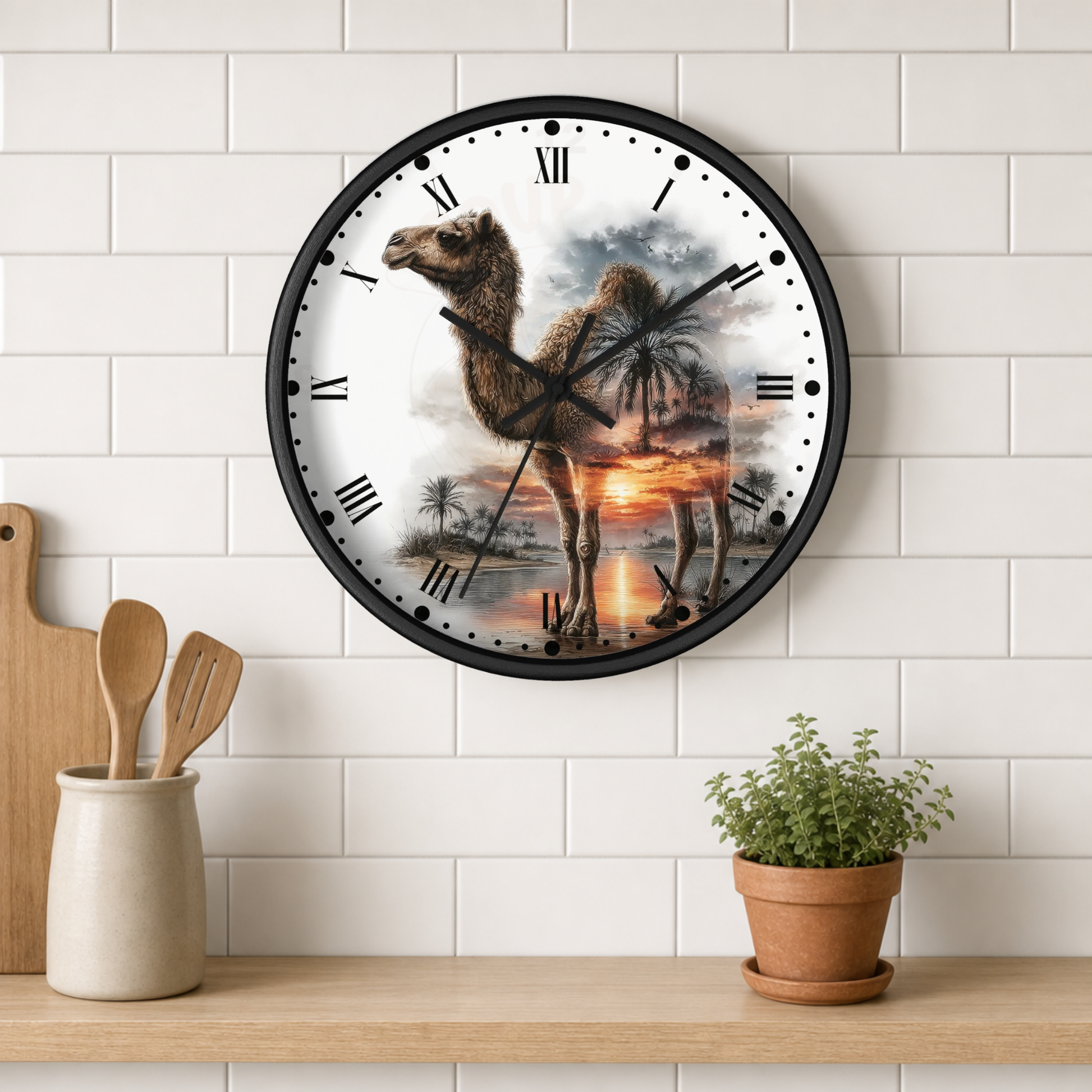 A round wall clock with a black frame hangs on a white subway tile wall above a wooden shelf. The clock face features a printed desert scene with a camel, palm trees, and a sunset.