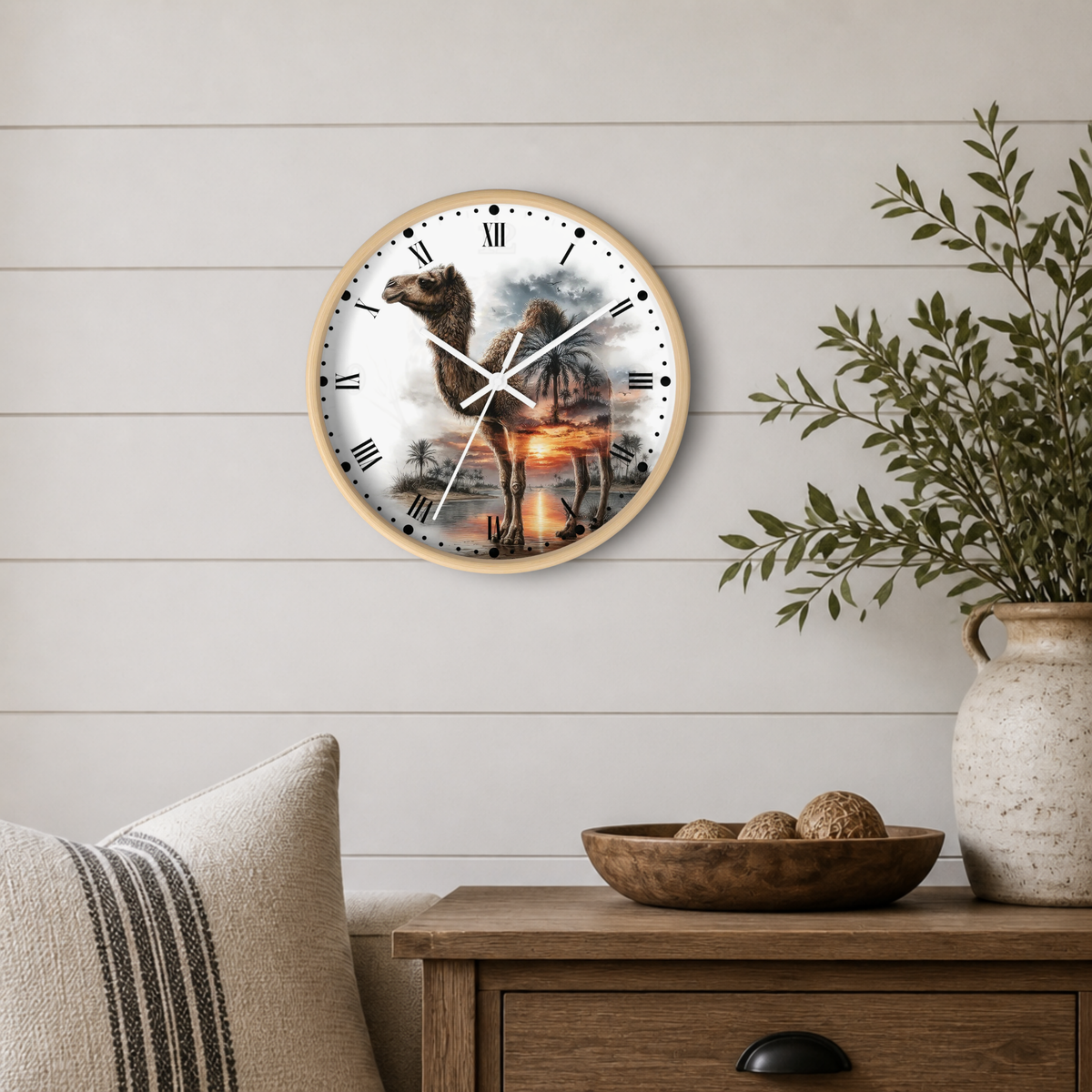 Round wall clock with a camel illustration, mounted on a white shiplap wall above a wooden side table with a vase and bowl.
