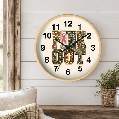 A round wall clock with a light wood frame hangs on a white shiplap wall above a wooden table. The clock face is cream with black numerals and hands, featuring the words 'PINK OUT' in camouflage block letters, with a pink awareness ribbon over the letter O.