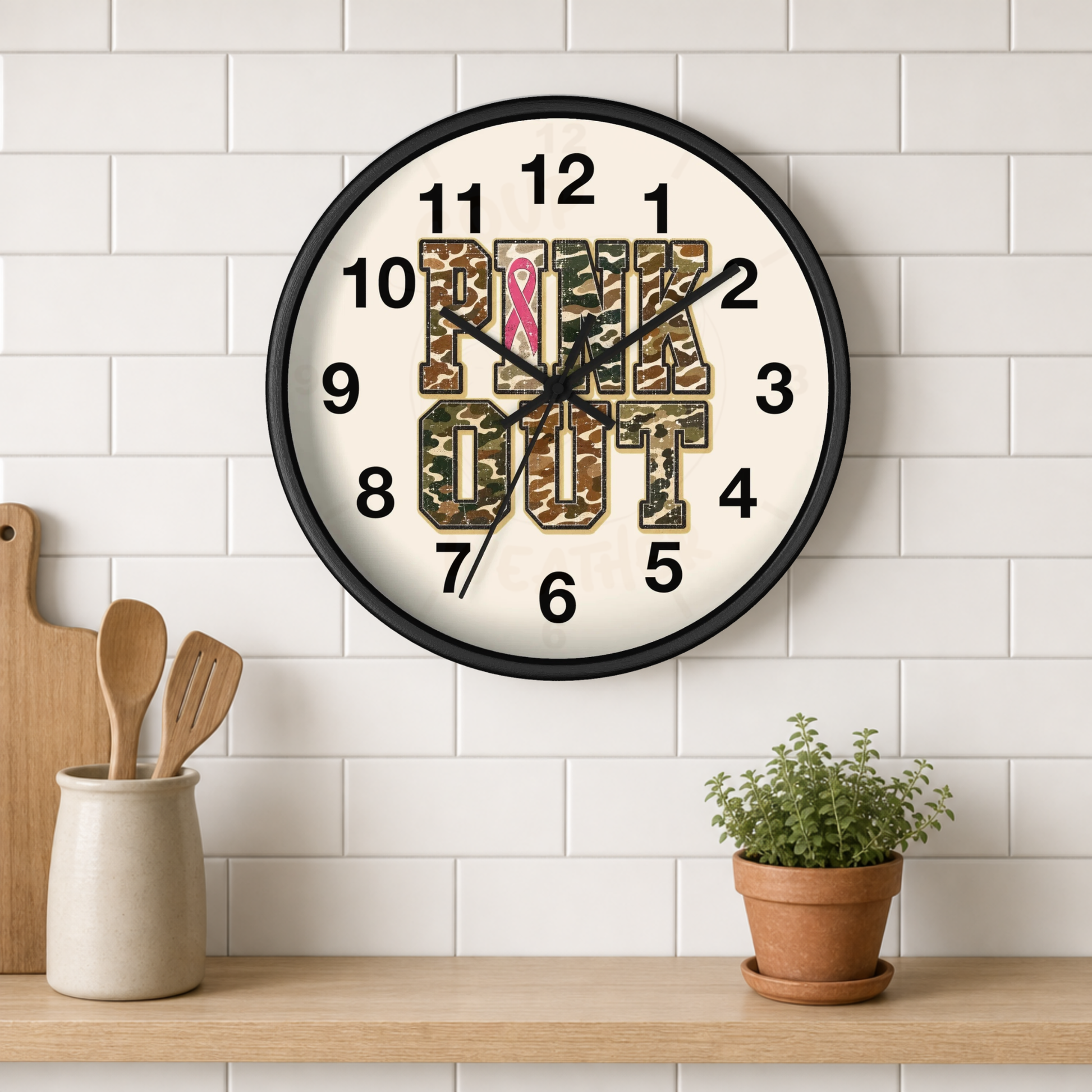 A round wall clock with a black frame hangs on a white subway tile wall above a wooden shelf. The clock face is white with black numerals and hands, featuring the words 'Pink OUT' in camouflage lettering with a pink ribbon symbol at the 12 position.