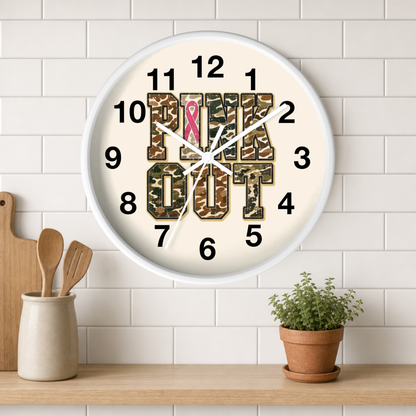 A round wall clock with a white frame hangs on a white subway tile wall above a wooden shelf. The clock face is cream-colored with bold black numbers and the words 'PINK OUT' in camouflage block letters, with a pink ribbon at the 12 o'clock position.