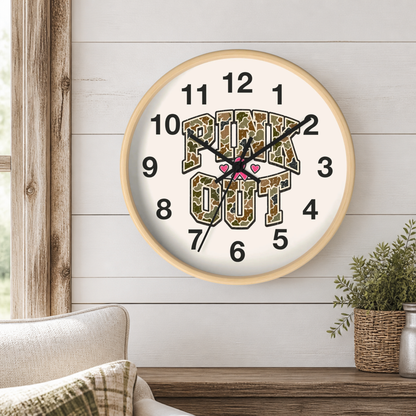 A round wall clock with a light wood frame hangs on a white shiplap wall above a wooden table. The clock face is white with black numerals and hands, featuring the words 'Pink OUT' in a camouflage pattern with small pink hearts in the center.