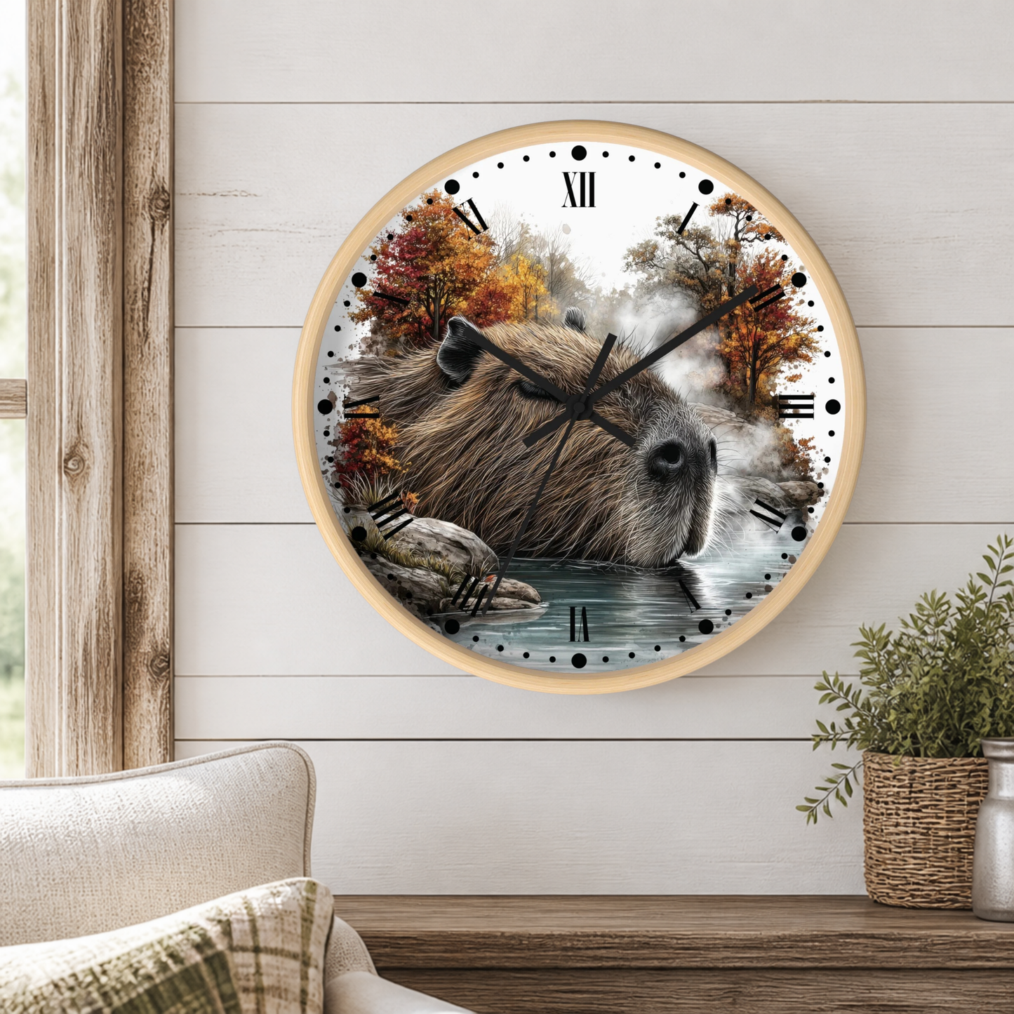 A round wall clock with a light wood frame hangs on a white shiplap wall above a wooden table. The clock face features a detailed illustration of a capybara sitting in shallow water, surrounded by autumn trees and mist.