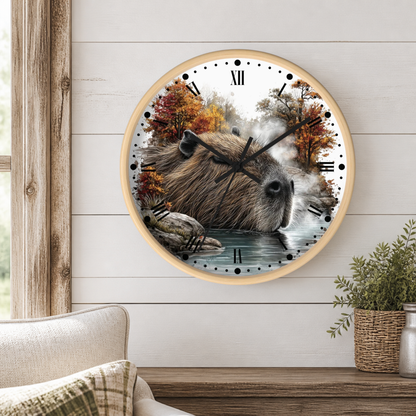A round wall clock with a light wood frame hangs on a white shiplap wall above a wooden table. The clock face features a detailed illustration of a capybara sitting in shallow water, surrounded by autumn trees and mist.
