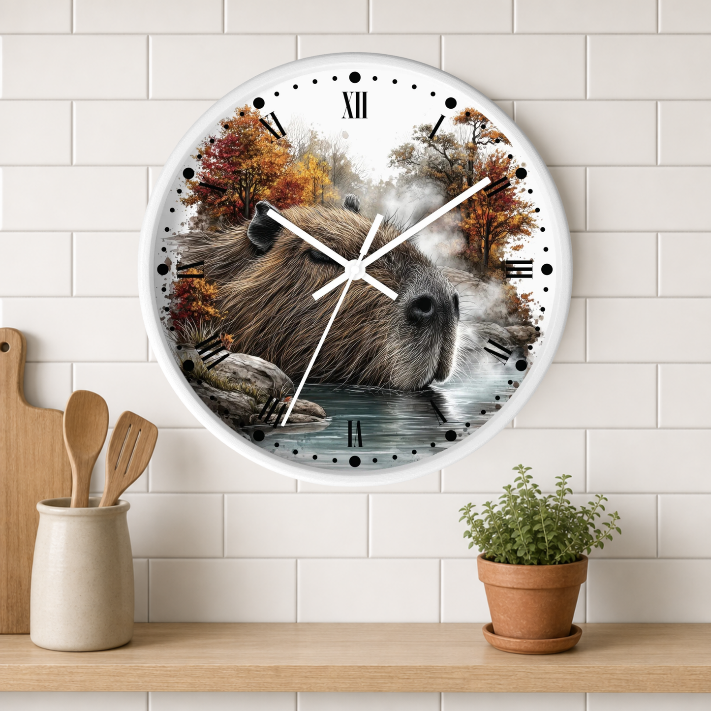 A round wall clock with a white frame and Roman numerals, featuring a printed design of a capybara in a forest by water. The clock is mounted on a white subway tile wall above a wooden shelf with kitchen utensils and a small potted plant.