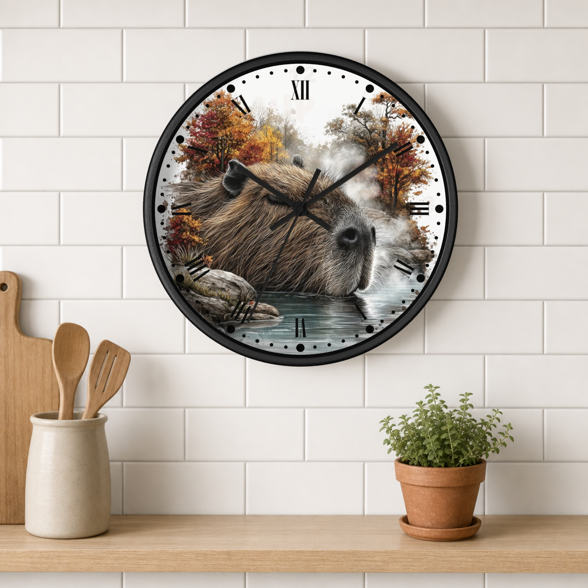 A round wall clock with a black frame hangs on a white subway tile wall above a wooden shelf. The clock face features a detailed illustration of a capybara in an autumn forest by a body of water.