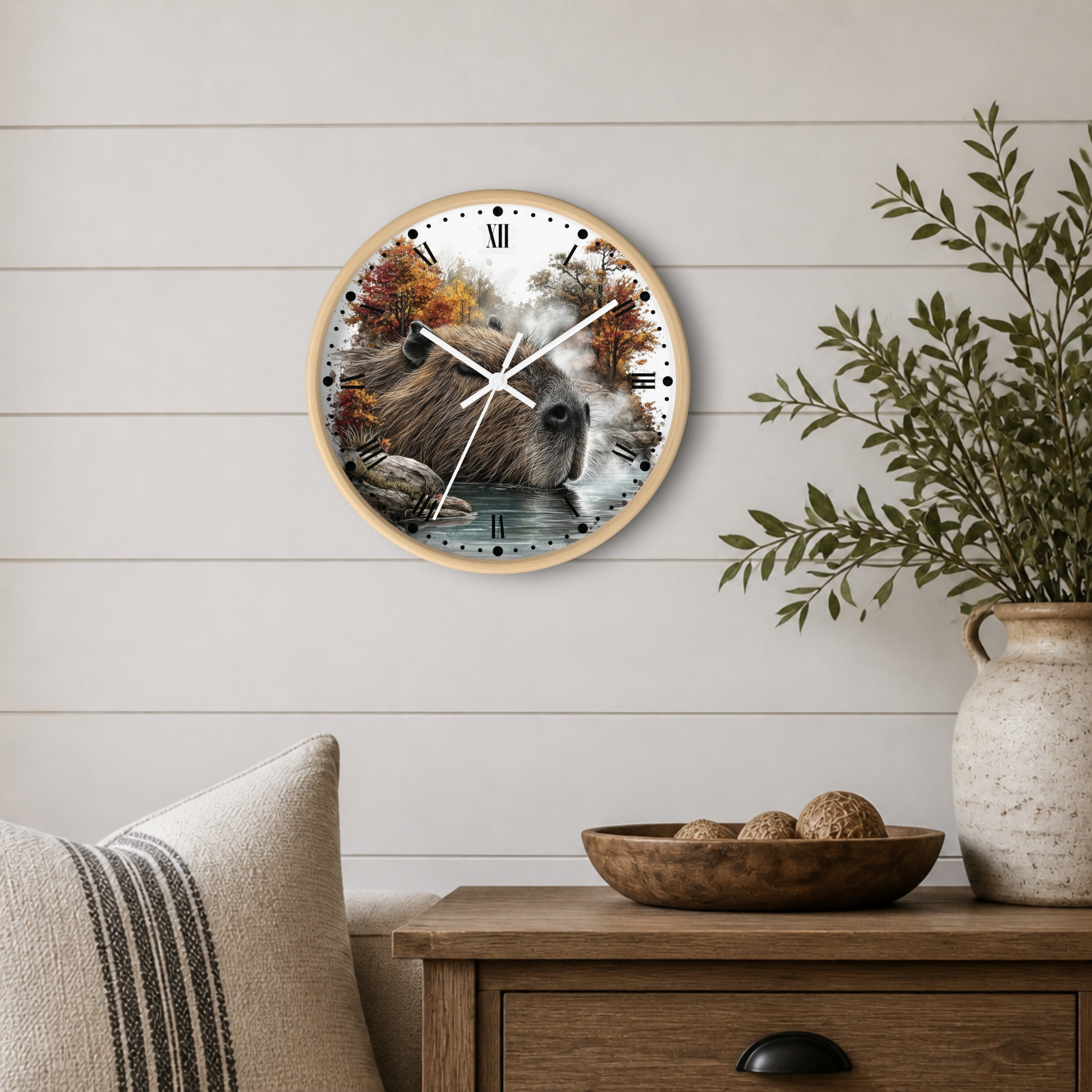 A round wall clock with a light wood frame hangs on a white shiplap wall above a wooden side table. The clock face features a detailed illustration of a capybara in a forest with autumn trees and water.