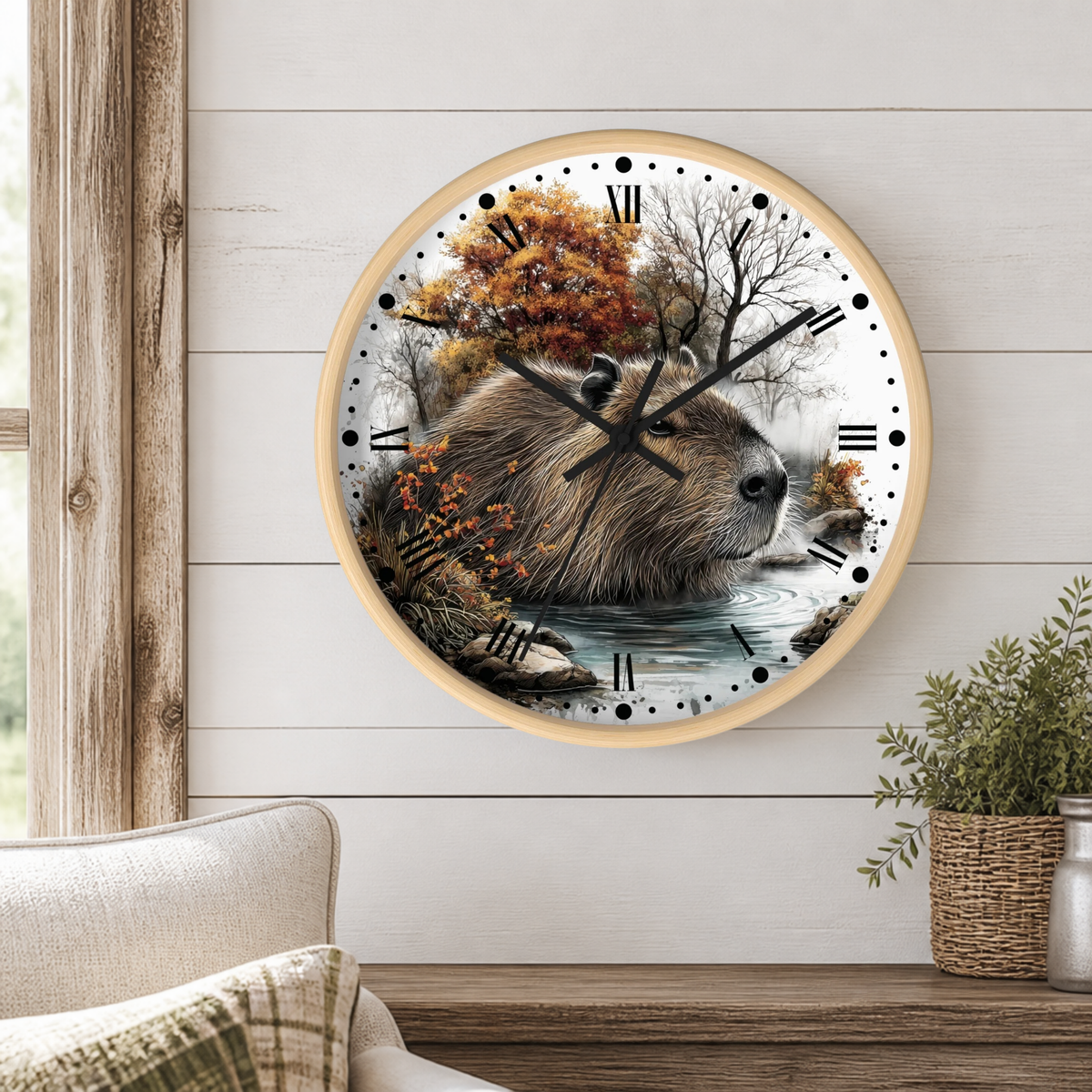 A round wall clock with a light wood frame hangs on a white shiplap wall above a wooden table. The clock face features a detailed illustration of a capybara surrounded by autumn trees and water, with black Roman numerals and hands.