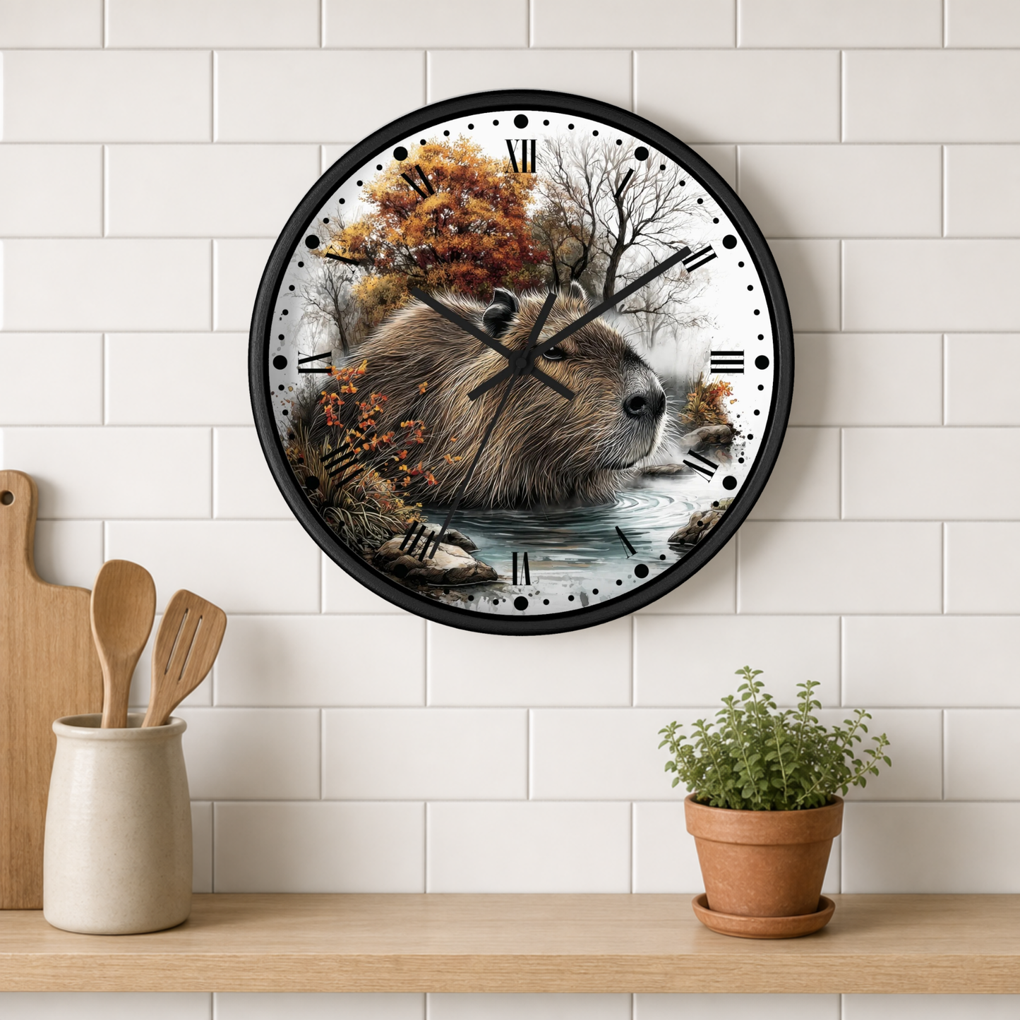 A round wall clock with a black frame hangs on a white subway-tile wall above a wooden shelf. The clock face features a detailed illustration of a capybara in a river surrounded by autumn trees.