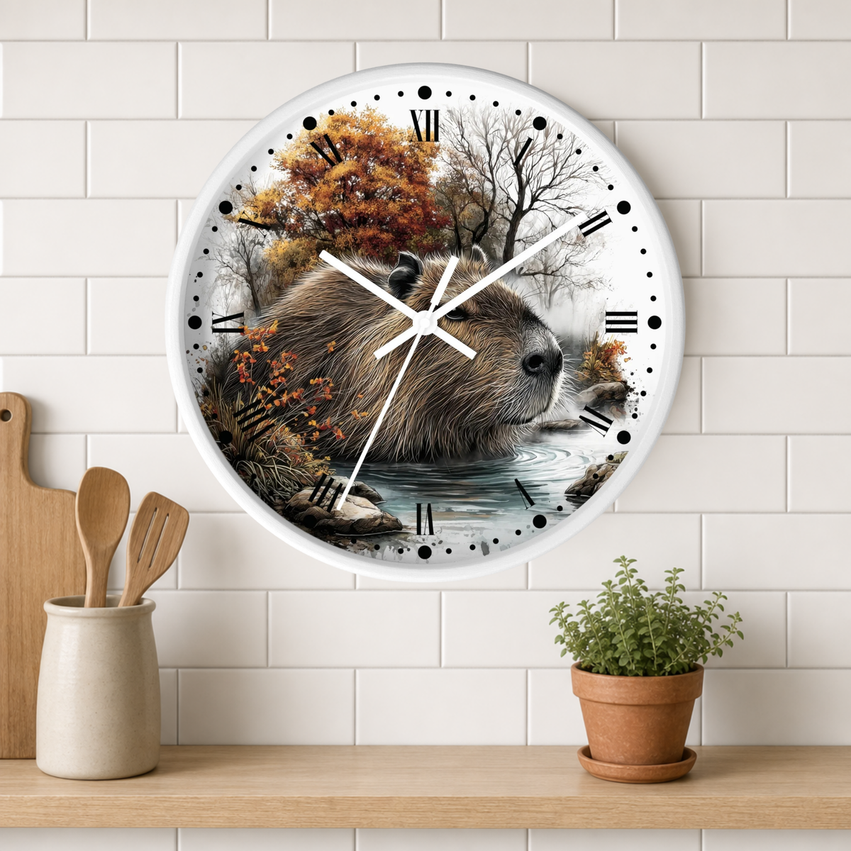 A round wall clock with a white frame hangs on a white subway-tile wall above a wooden shelf. The clock face features a detailed illustration of a capybara in an autumn forest by a river.