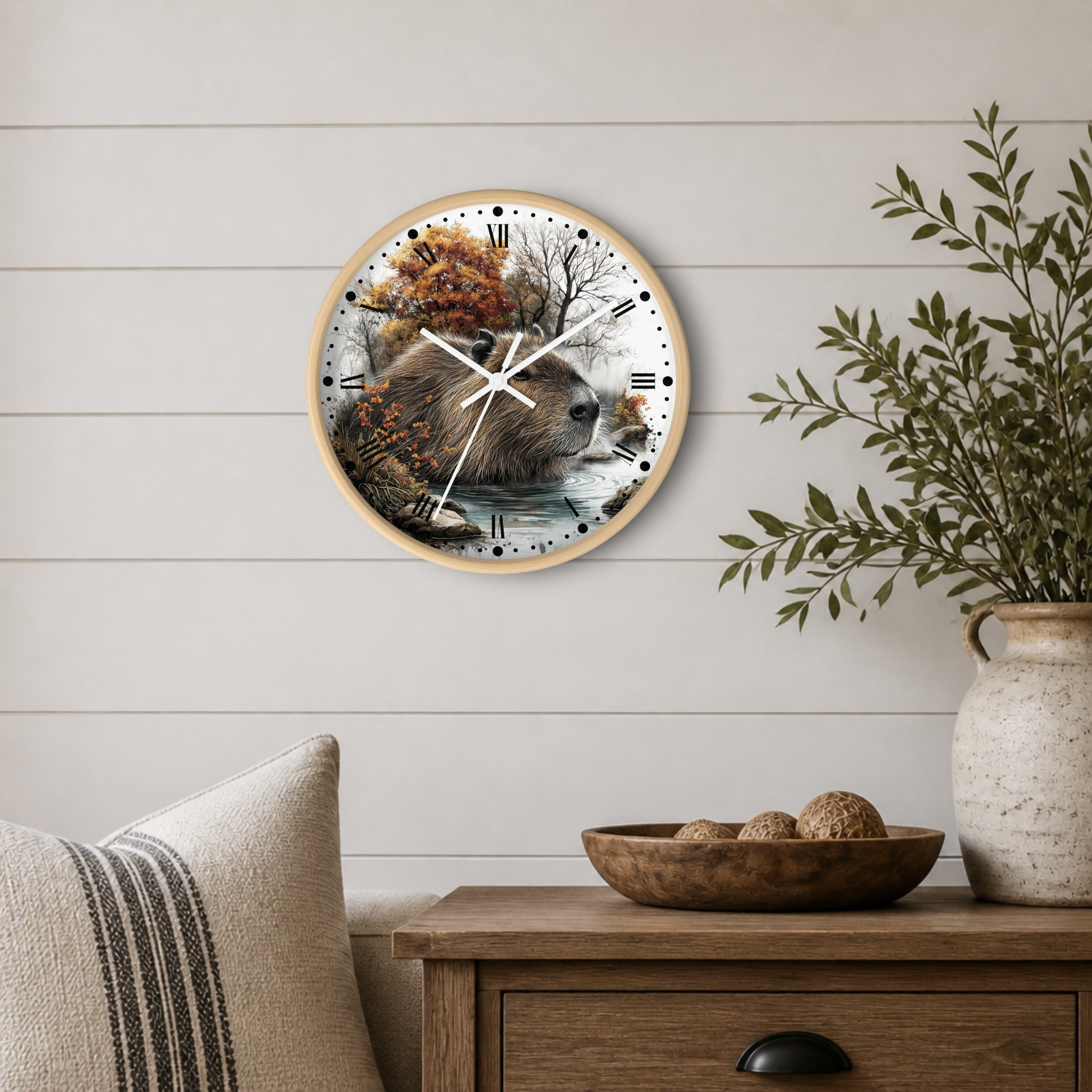 Round wall clock with a natural wood frame and a detailed capybara illustration on the face, styled on a white shiplap wall above a wooden side table with a vase and bowl.