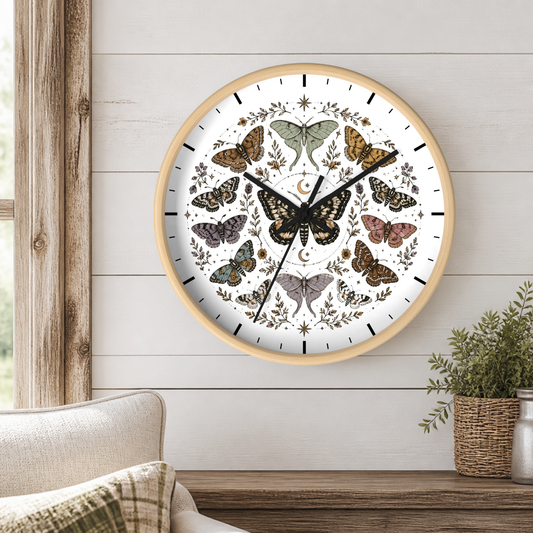 A round wall clock with a light wood frame and a white face featuring multiple illustrated butterflies and botanical motifs, mounted on a white shiplap wall above a wooden table with a potted plant.