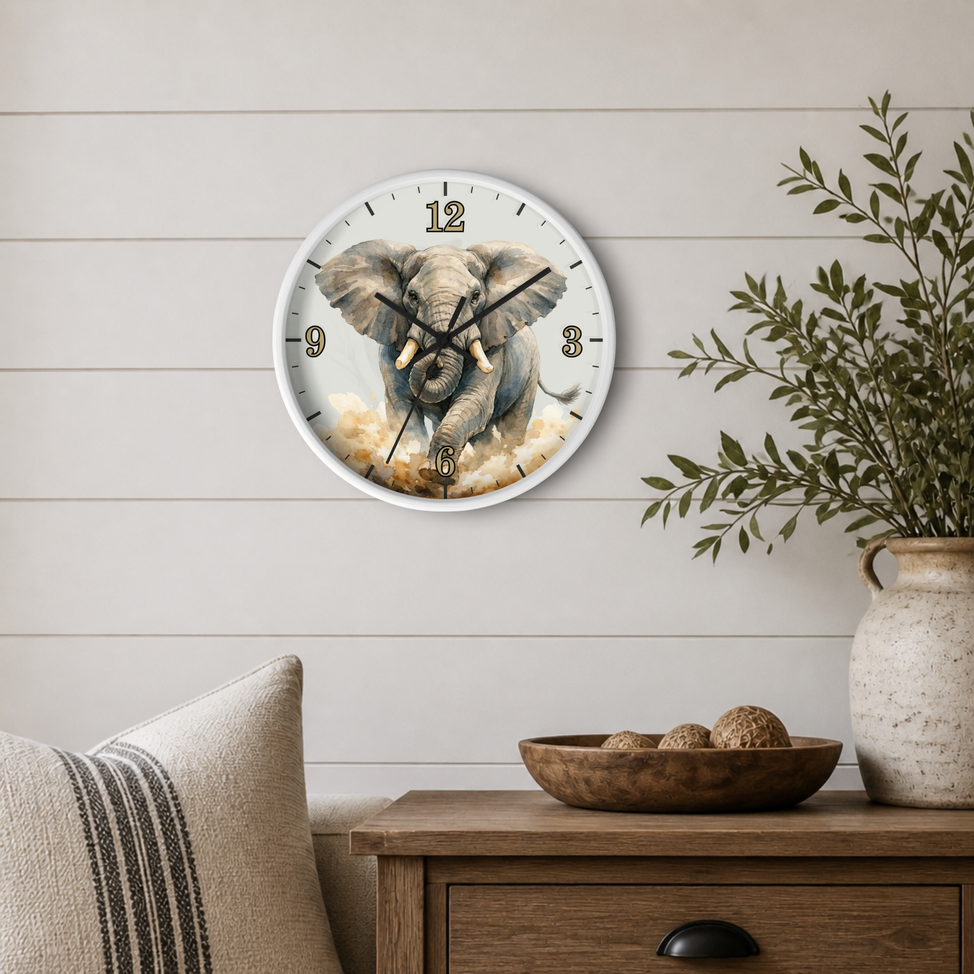 A round wall clock featuring a painted elephant illustration, mounted on a light wood shiplap wall above a wooden side table with a bowl and a potted plant.