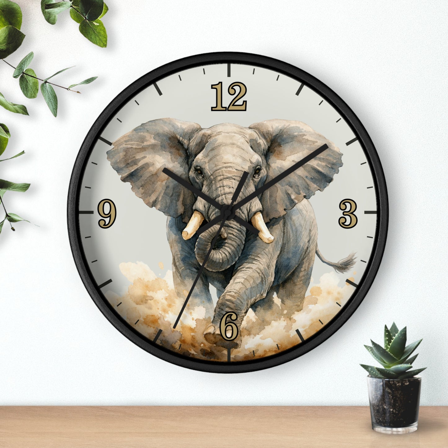 A round wall clock with a black frame and a printed elephant illustration on the face, shown hanging on a white wall above a wooden surface with small green plants nearby.