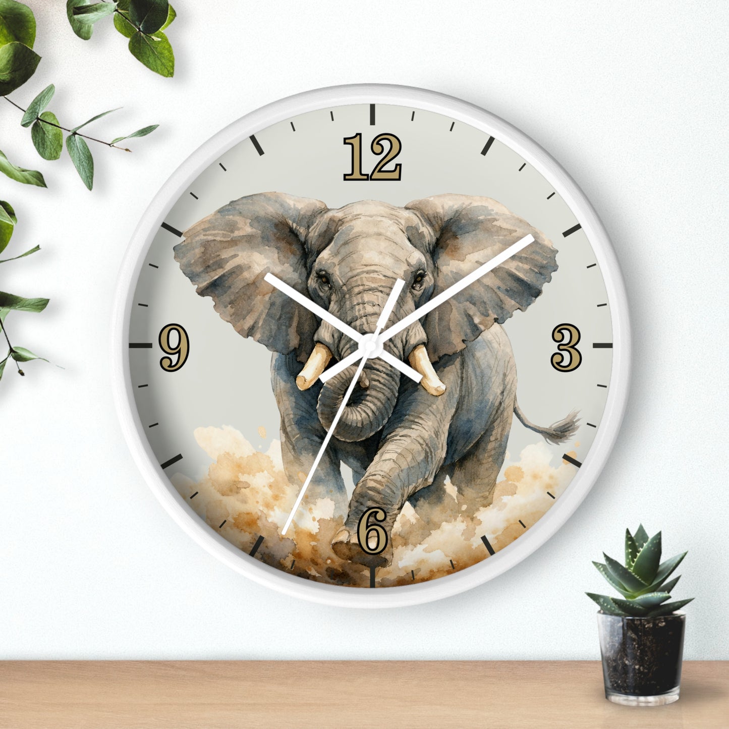 A round wall clock with a white frame and a printed elephant illustration on the face, shown hanging on a white wall above a wooden surface with small plants nearby.