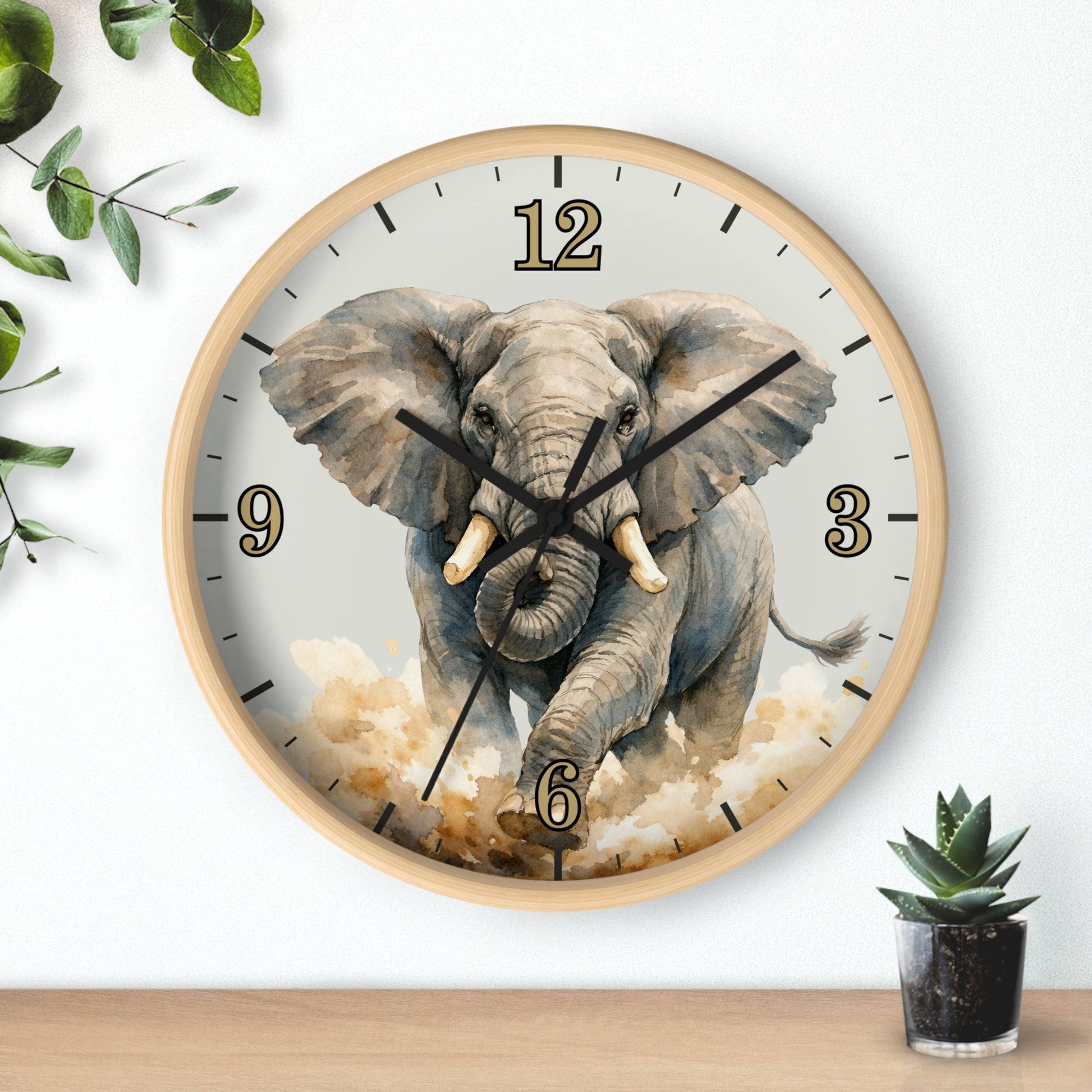 Round wall clock with a light wood frame and a printed elephant design on the face, shown hanging on a white wall above a wooden surface with small green plants nearby.