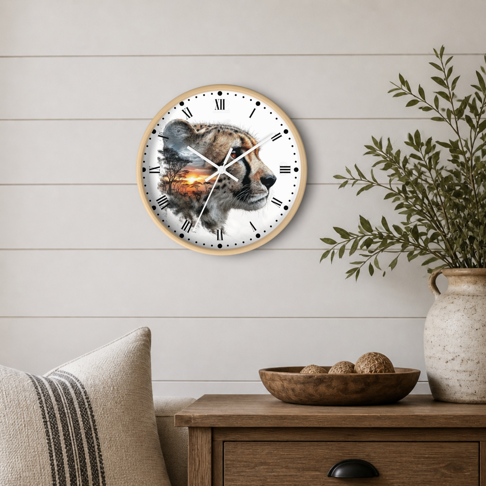 A round wall clock with a light wood frame and a white face featuring a detailed cheetah illustration with a warm orange sunset glow in the center, mounted on a white shiplap wall above a wooden side table with a plant and decorative bowl.
