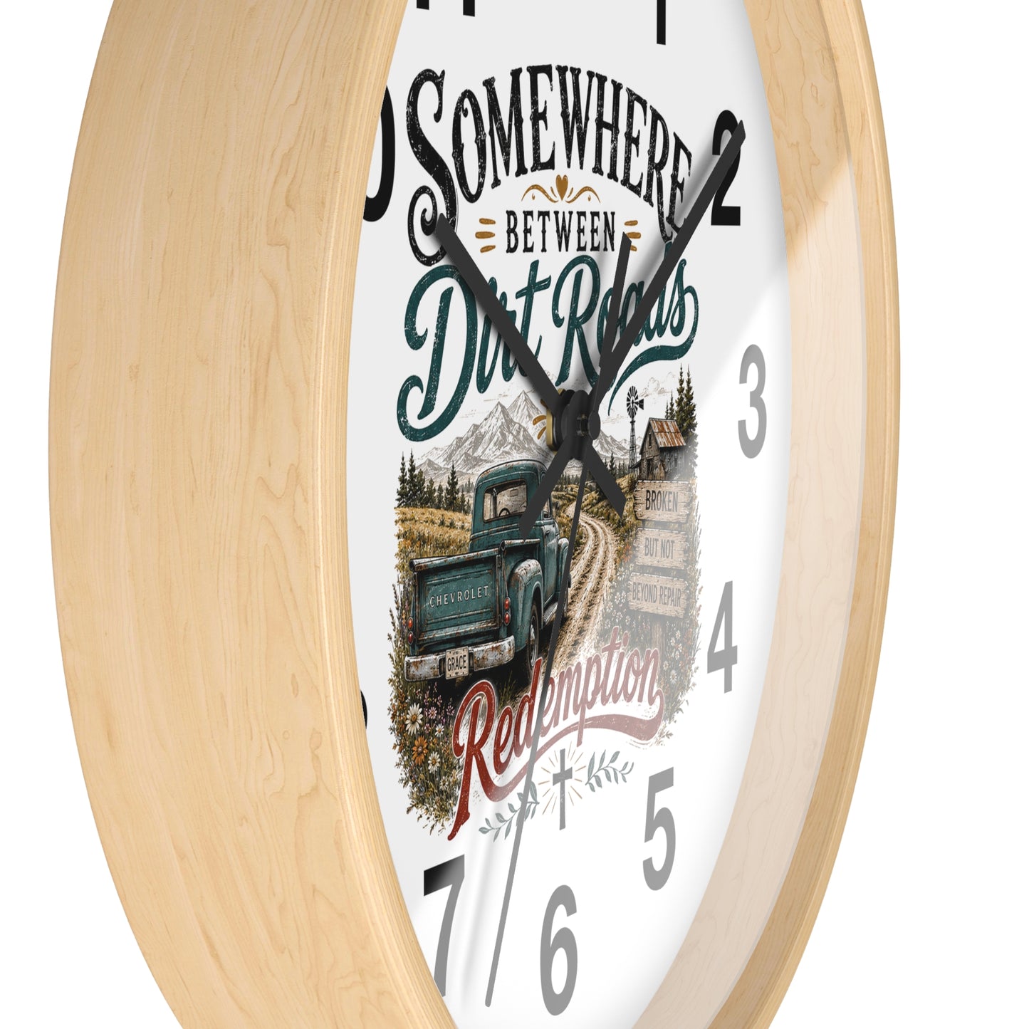 A round wall clock with a light natural wood frame and a white face featuring a rustic illustration of a green pickup truck on a dirt road, mountains, and the text 'Somewhere Between Dirt Roads Redemption'.