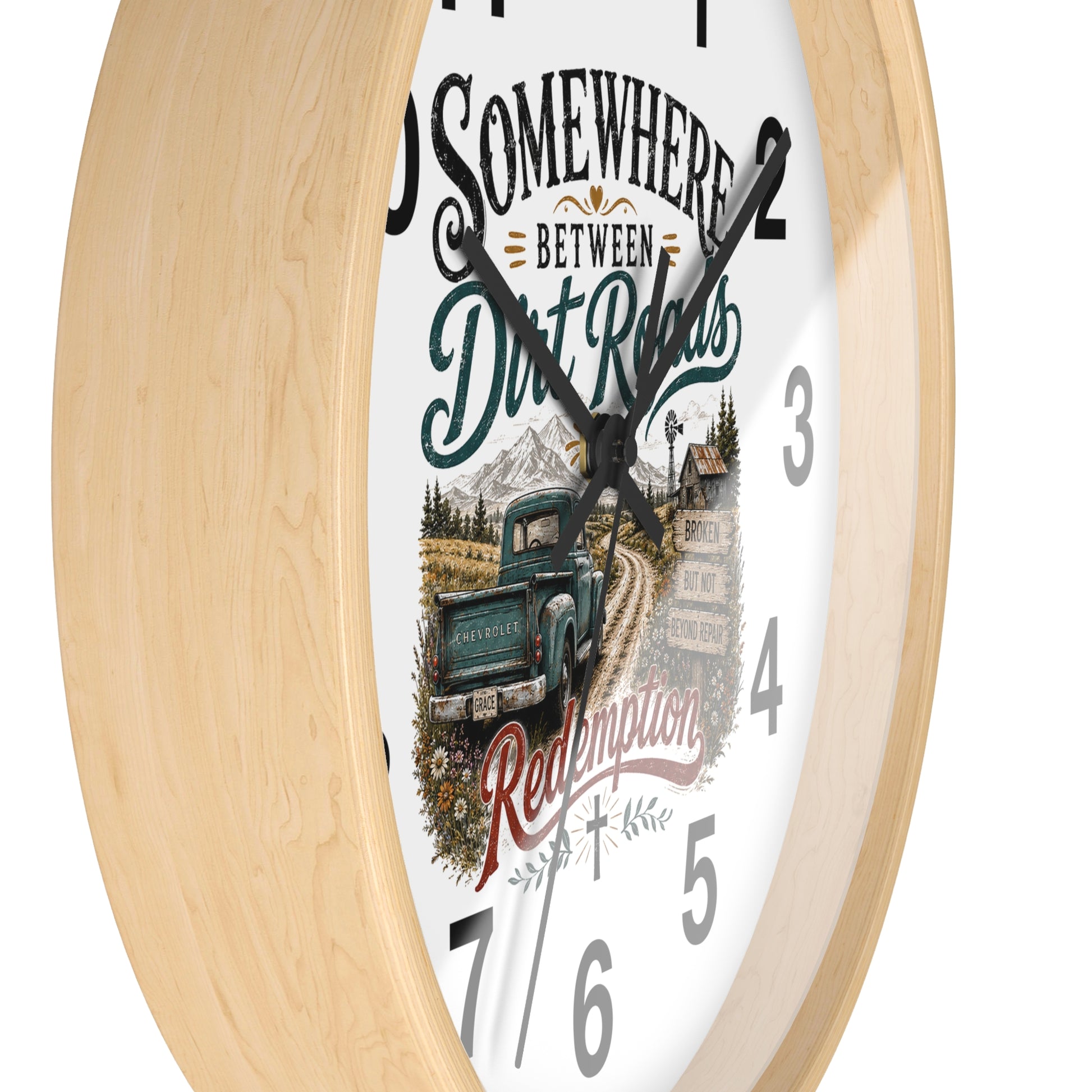 A round wall clock with a light natural wood frame and a white face featuring a rustic illustration of a green pickup truck on a dirt road, mountains, and the text 'Somewhere Between Dirt Roads Redemption'.