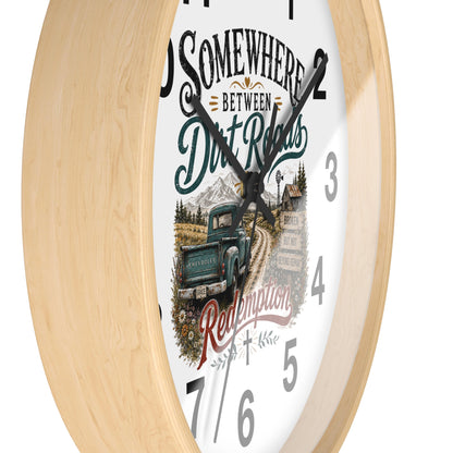 A round wall clock with a light natural wood frame and a white face featuring a rustic illustration of a green pickup truck on a dirt road, mountains, and the text 'Somewhere Between Dirt Roads Redemption'.