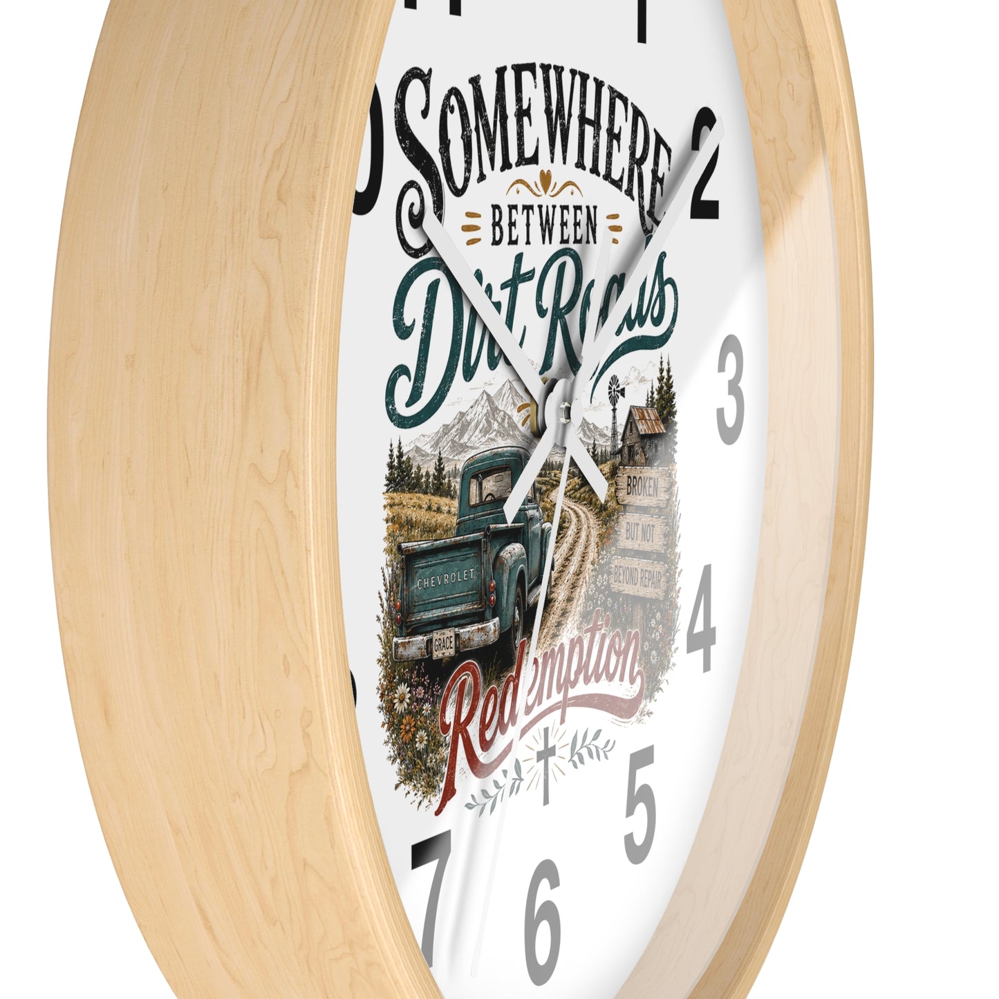 A round wall clock with a light natural wood frame and a white face featuring a rustic graphic design. The design includes the text 'SOMEWHERE BETWEEN Dirt Roads Red Emporia' in stylized fonts, along with an illustration of a vintage pickup truck on a dirt road in front of mountains and a cabin.
