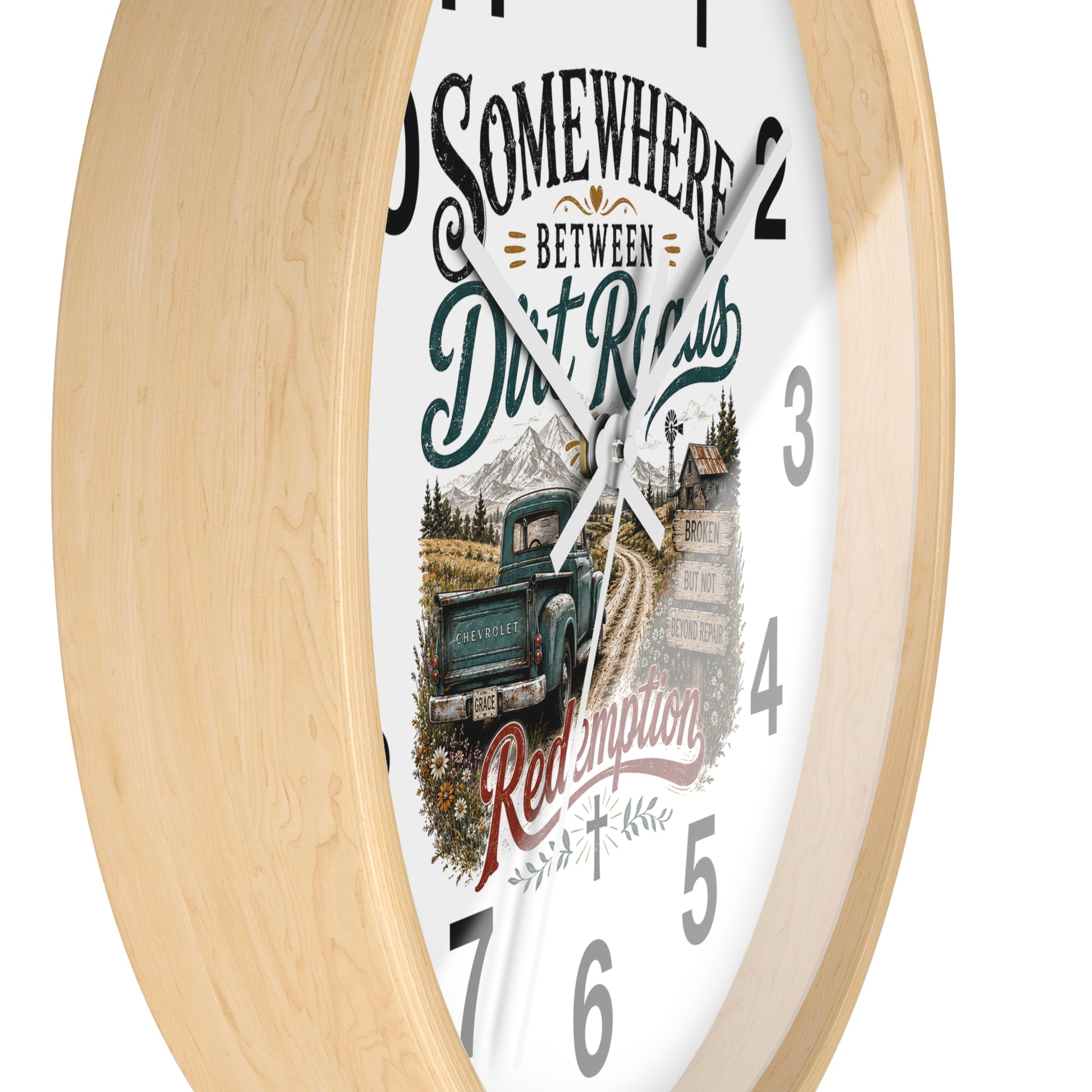 A round wall clock with a light natural wood frame and a white face featuring a rustic graphic design. The design includes the text 'SOMEWHERE BETWEEN Dirt Roads Red Emporia' in stylized fonts, along with an illustration of a vintage pickup truck on a dirt road in front of mountains and a cabin.