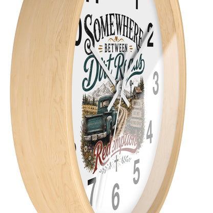 A round wall clock with a light natural wood frame and a white face featuring a rustic graphic design. The design includes the text 'SOMEWHERE BETWEEN Dirt Roads Red Emporia' in stylized fonts, along with an illustration of a vintage pickup truck on a dirt road in front of mountains and a cabin.