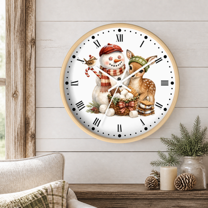 A round wall clock with a light wood frame hangs on a white shiplap wall above a rustic table with candles and pinecones. The clock face features a winter illustration of a snowman in a red hat and scarf, a deer, a bird, and a basket of marshmallows.
