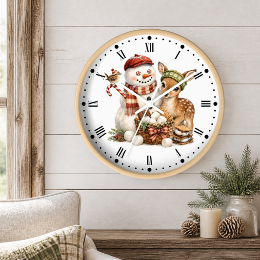 A round wall clock with a light wood frame hangs on a white shiplap wall above a rustic table with candles and pinecones. The clock face features a winter illustration of a snowman in a red hat and scarf, a deer, a bird, and a basket of marshmallows.