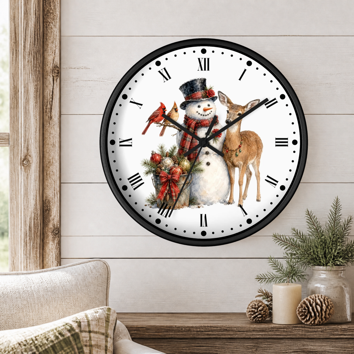 A round wall clock with a black frame and a festive winter illustration of a snowman, a deer, red cardinals, and holiday greenery on the dial, shown mounted on a light wood paneled wall above a table with candles and pinecones.