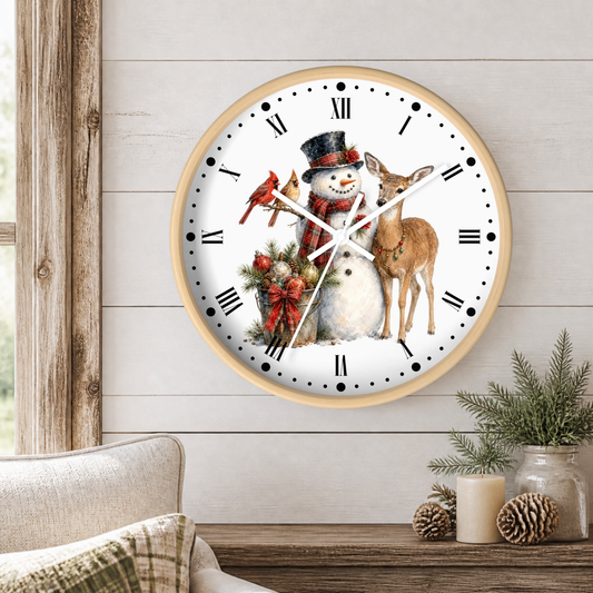 A round wall clock with a light wood frame hangs on a white shiplap wall above a rustic wooden table. The clock face features a winter illustration with a snowman in a black top hat and red scarf, a brown deer, a red cardinal, and a holiday wreath.