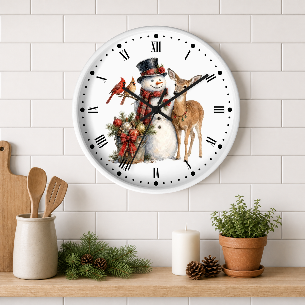 A round wall clock with a white face and black Roman numerals, decorated with a winter holiday illustration of a snowman, a deer, a red bird, and a small tree. The clock is mounted on a white subway tile wall above a wooden shelf styled with kitchen utensils, pine branches, a candle, and a potted plant.